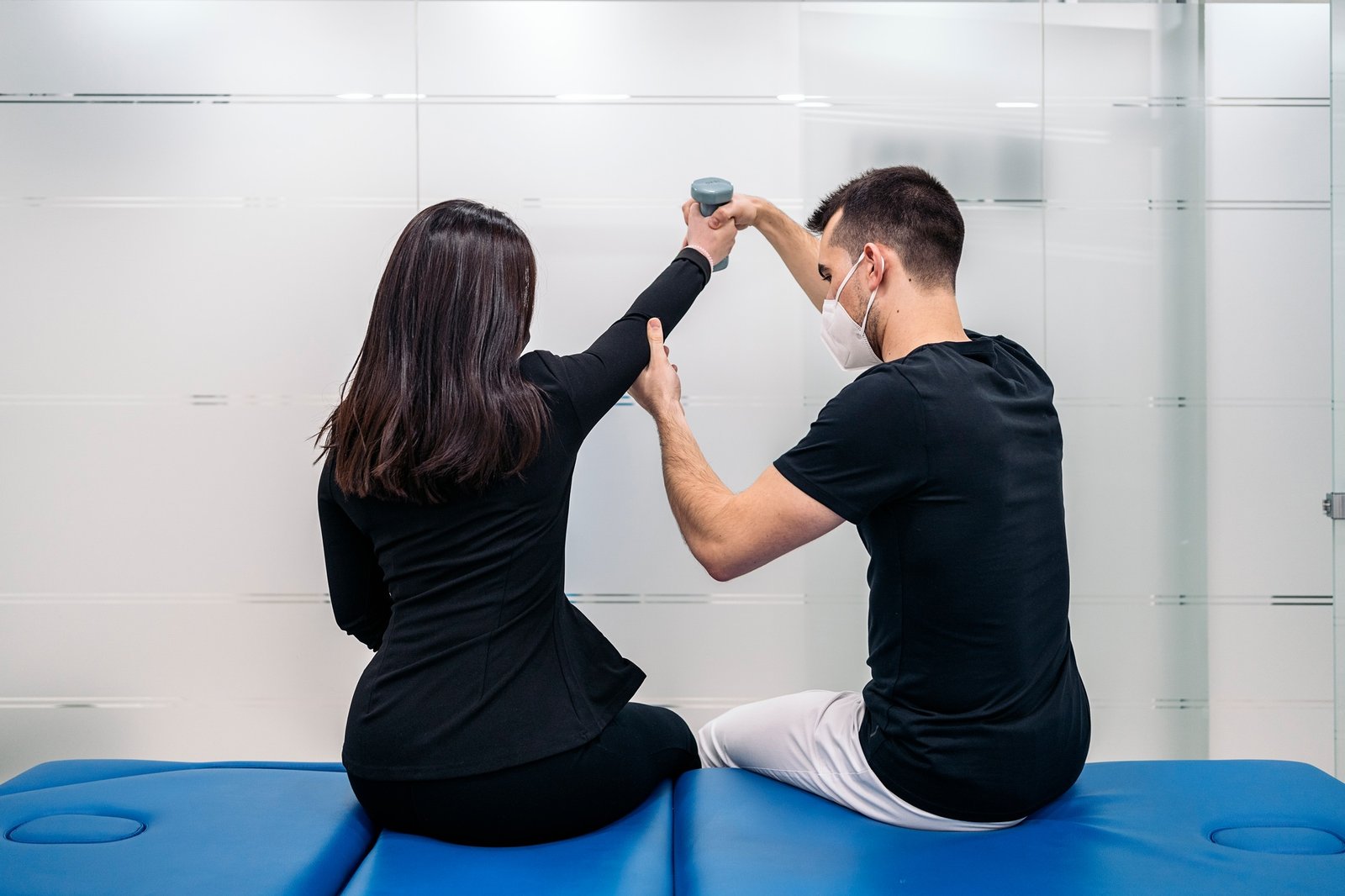 Fisioterapeuta asistiendo a una mujer a levantar el brazo con una pesa pequeña, ambos sentados sobre una camilla azul.